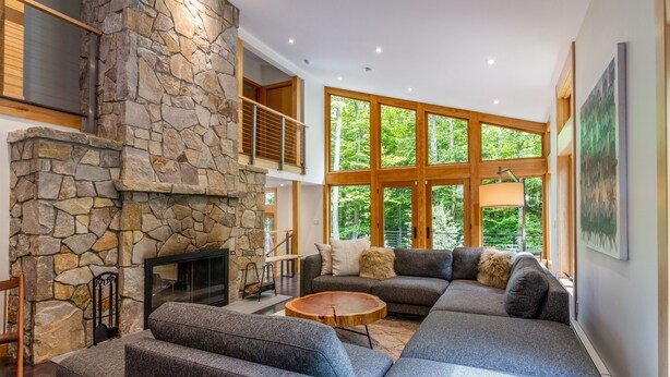 A modern living room with a stone fireplace, large windows, and a sectional sofa. - Luxurious Modern Home Steps to Gondola & Village (Stratton)