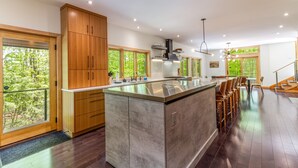 A modern kitchen with a large island, wooden cabinets, and a view of the forest. - Luxurious Modern Home Steps to Gondola & Village (Stratton)