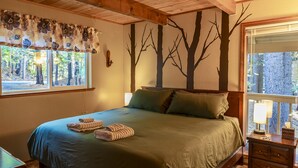 A neatly made bed with green bedding, a window with a floral curtain, and a wall with a tree mural. - Evergreen Dorrington • Both Cabins • Game Room • Hot Tub • Pool Access (Dorrington)