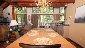 A spacious kitchen with a long wooden table, a chandelier, and a view of the outdoors through large windows. - Evergreen Dorrington • Both Cabins • Game Room • Hot Tub • Pool Access (Dorrington)