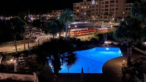 Vue nocturne d’un hôtel avec une piscine, des palmiers et une marina en arrière-plan.