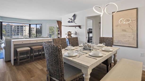 Un espace de repas moderne, avec une longue table dressée pour quatre personnes, entourée de chaises en osier. Une grande fenêtre offre une vue sur le paysage urbain.