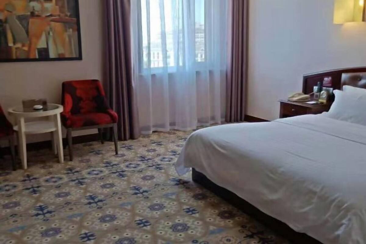 A hotel room with a bed, a chair, a small table, a nightstand, and a painting on the wall.