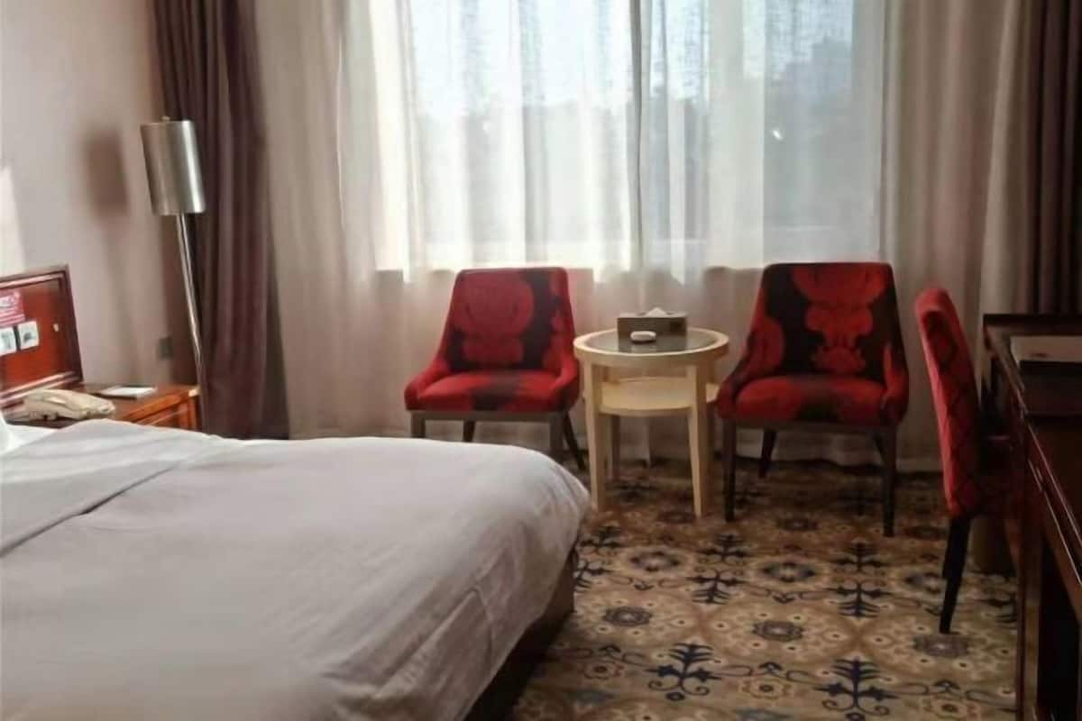 A hotel room with a bed, two red chairs, a small table, and a lamp.