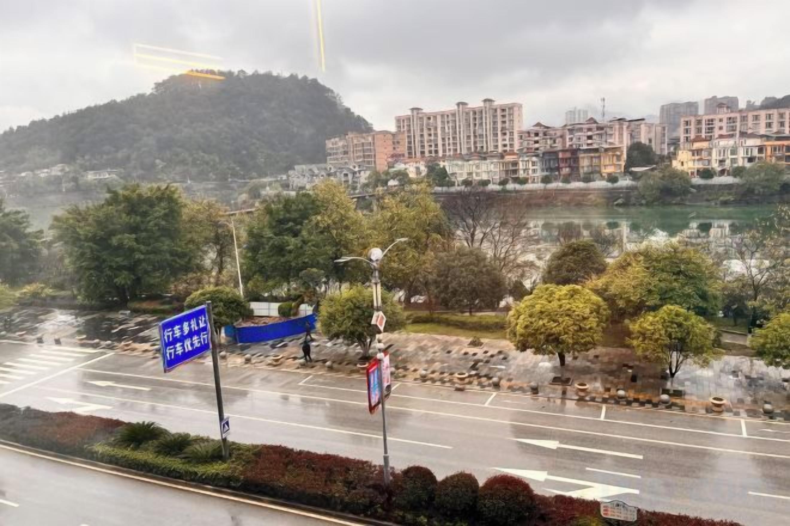 Una carretera con un cartel azul, árboles y edificios al fondo.