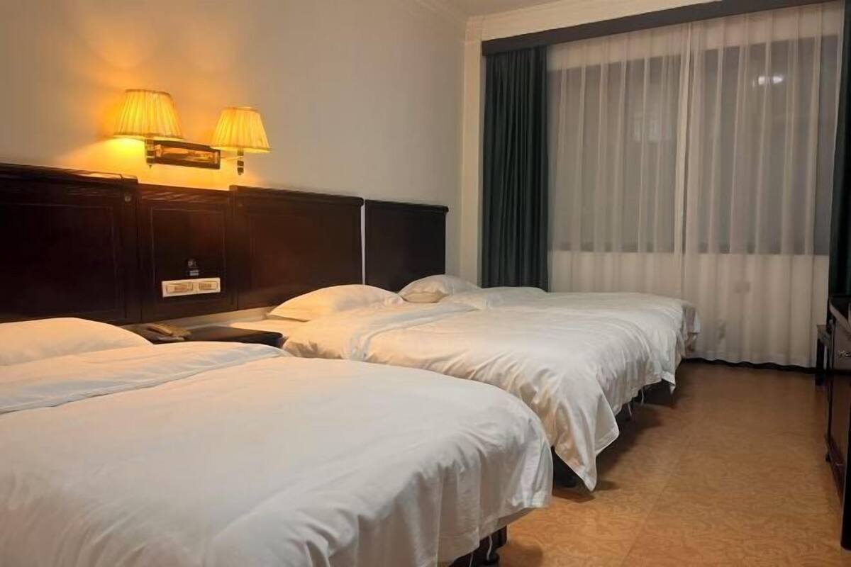 A hotel room with three single beds, a dark wooden headboard, a large window with sheer curtains, and wall-mounted lamps.
