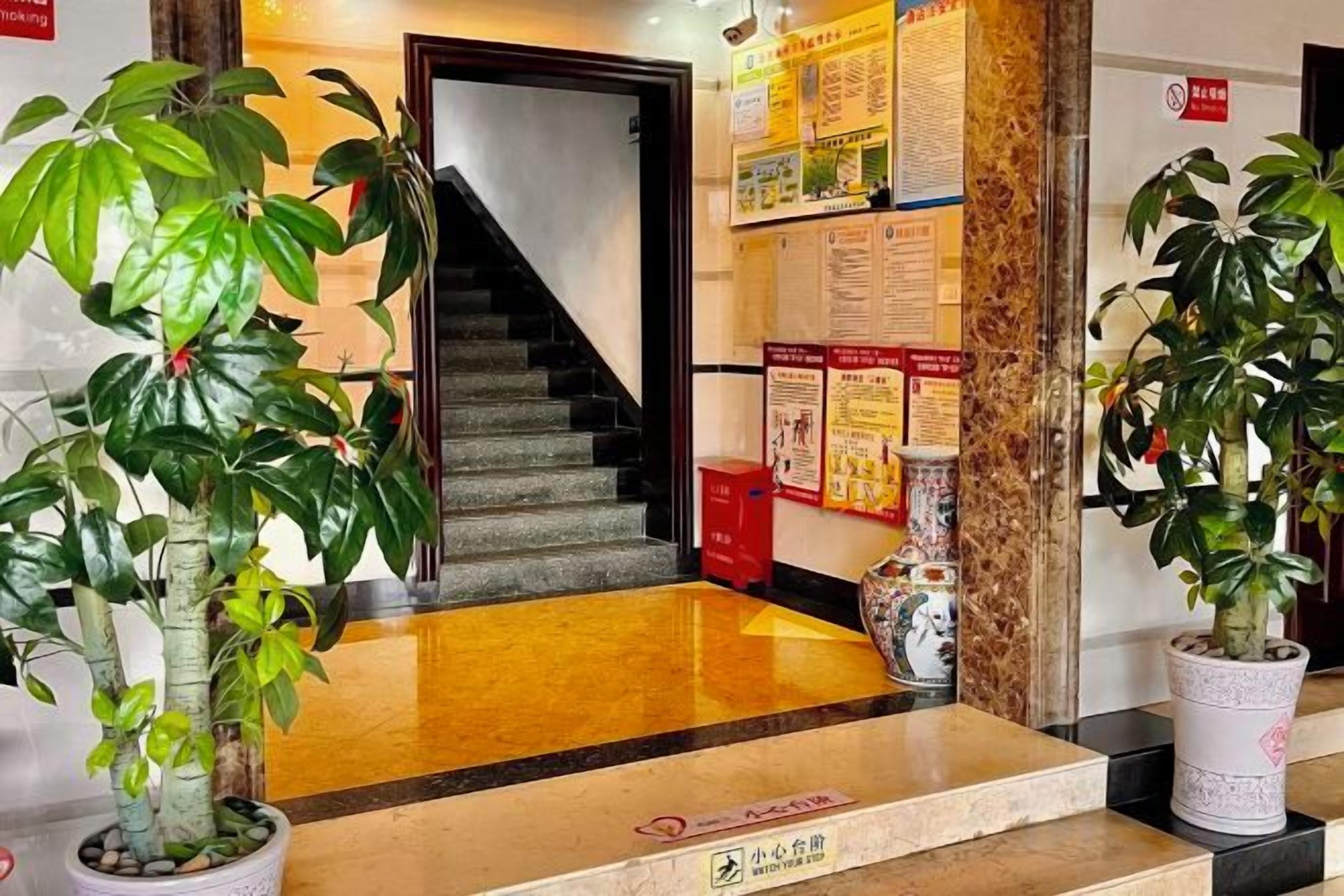 A hotel lobby with a staircase, potted plants, and informational posters on the walls.