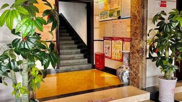 A hotel lobby with a staircase, potted plants, and informational posters on the walls.