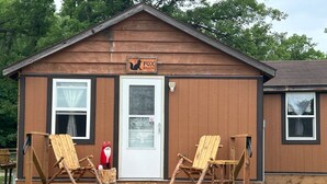 Una pequeña cabaña de madera con un letrero que dice "Fox Hollow". Tiene dos sillas de madera en un porche, una puerta y ventanas.
