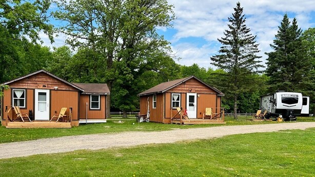 Dos cabañas de madera pequeñas con porches, cada una con puerta y ventanas, y una caravana estacionada cerca.