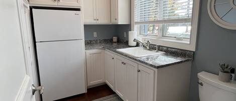 A compact kitchen with white cabinets, a refrigerator, a sink, and a window with blinds.