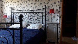 Um quarto com estrutura de cama em metal, colcha azul, almofadas brancas, abajur vermelho sobre uma mesa de cabeceira e um papel de parede com padrão floral.