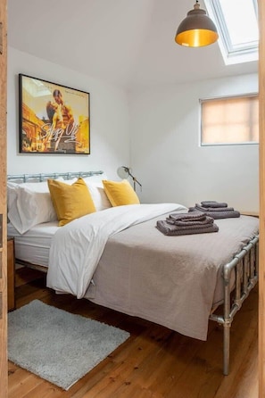 A bedroom with a bed, pillows, a bedside lamp, a framed picture on the wall, and a window with blinds.