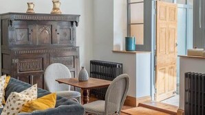 Quarto com sofá azul, mesa de jantar com cadeiras, armário de madeira escura e uma grande janela com vista externa.