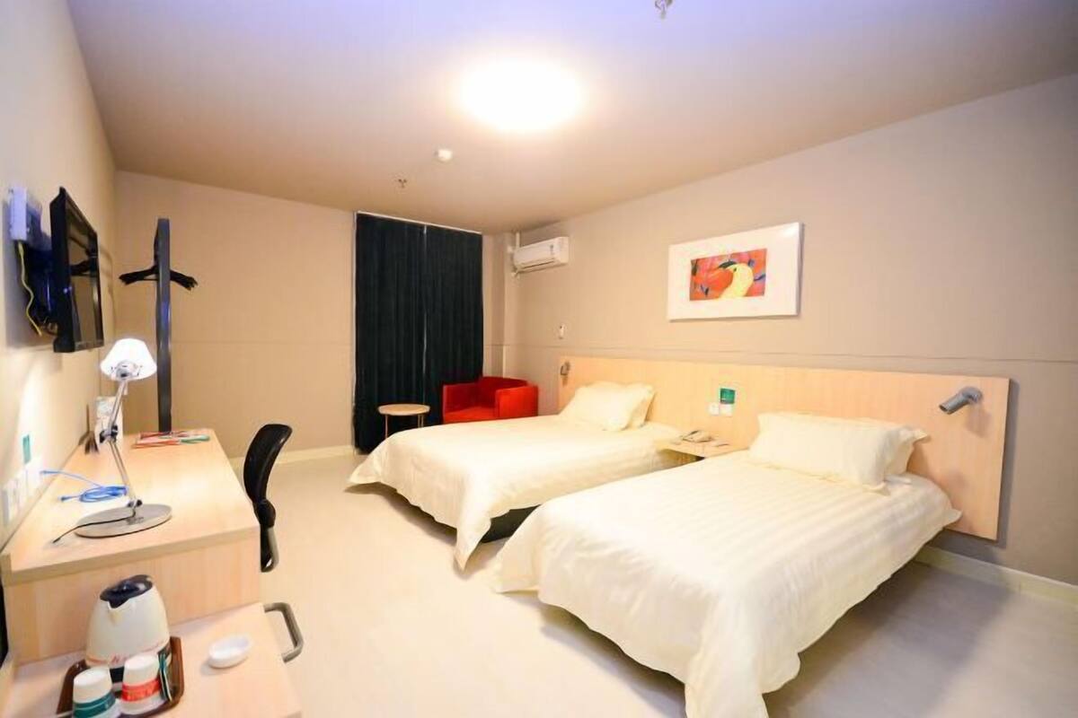 A hotel room with two beds, a desk with a lamp, a chair, a red armchair, and a painting on the wall.