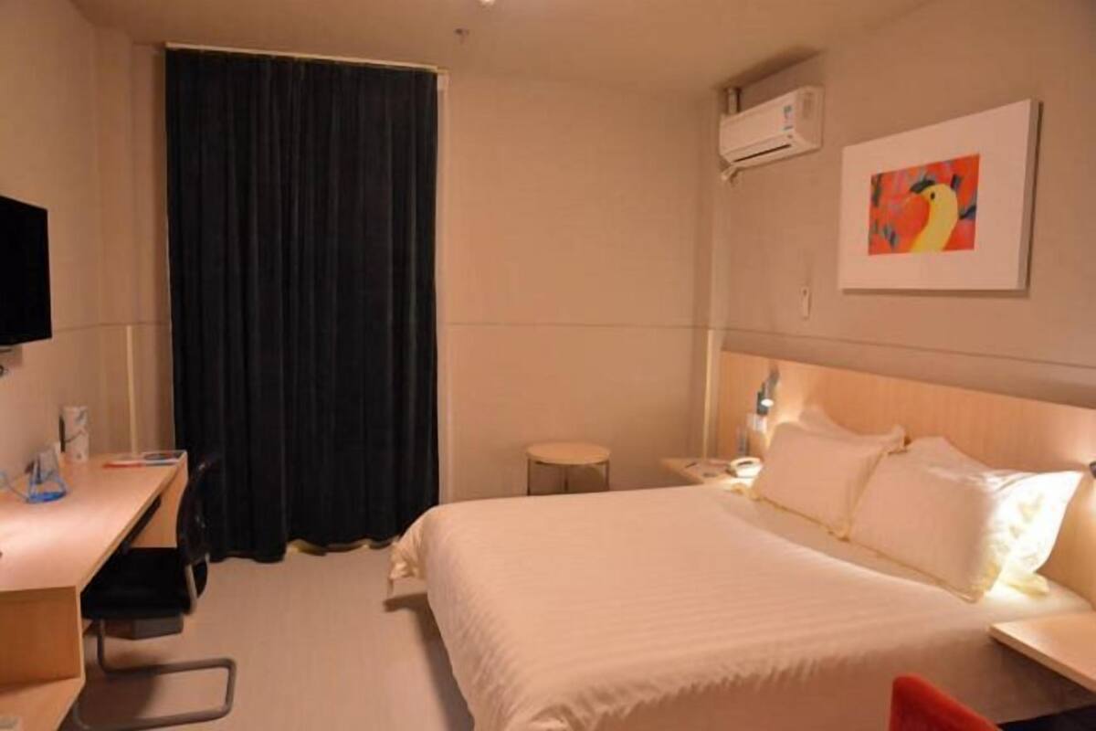 A hotel room with a bed, a desk, a chair, a small table, and a wall-mounted air conditioner.