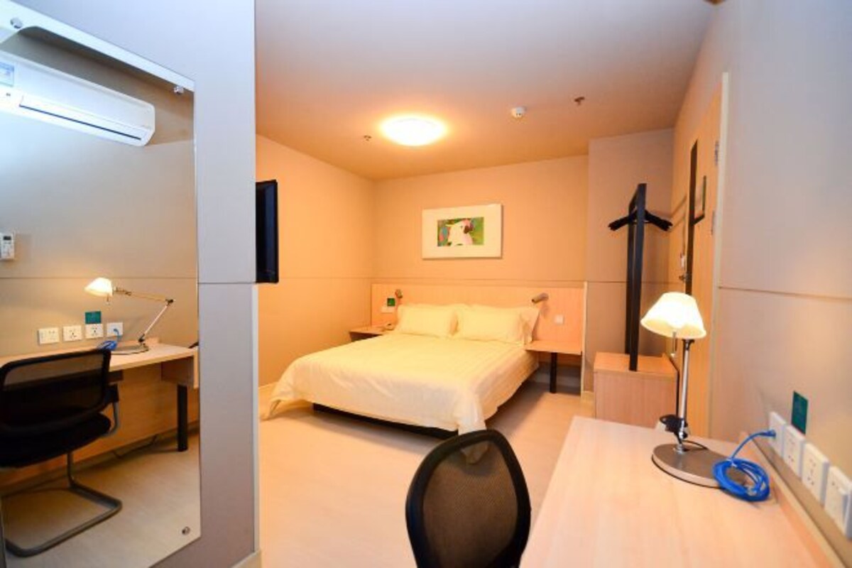 Ett hotellrum med en säng, ett skrivbord, en stol, en skrivbordslampa och en väggmonterad luftkonditioneringsanläggning.