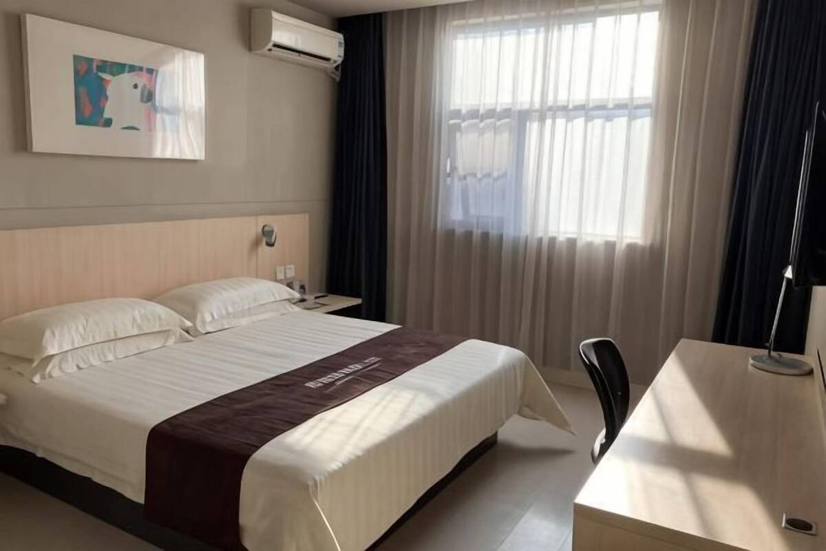 A hotel room with a bed, bedside table, chair, and a desk with a lamp.