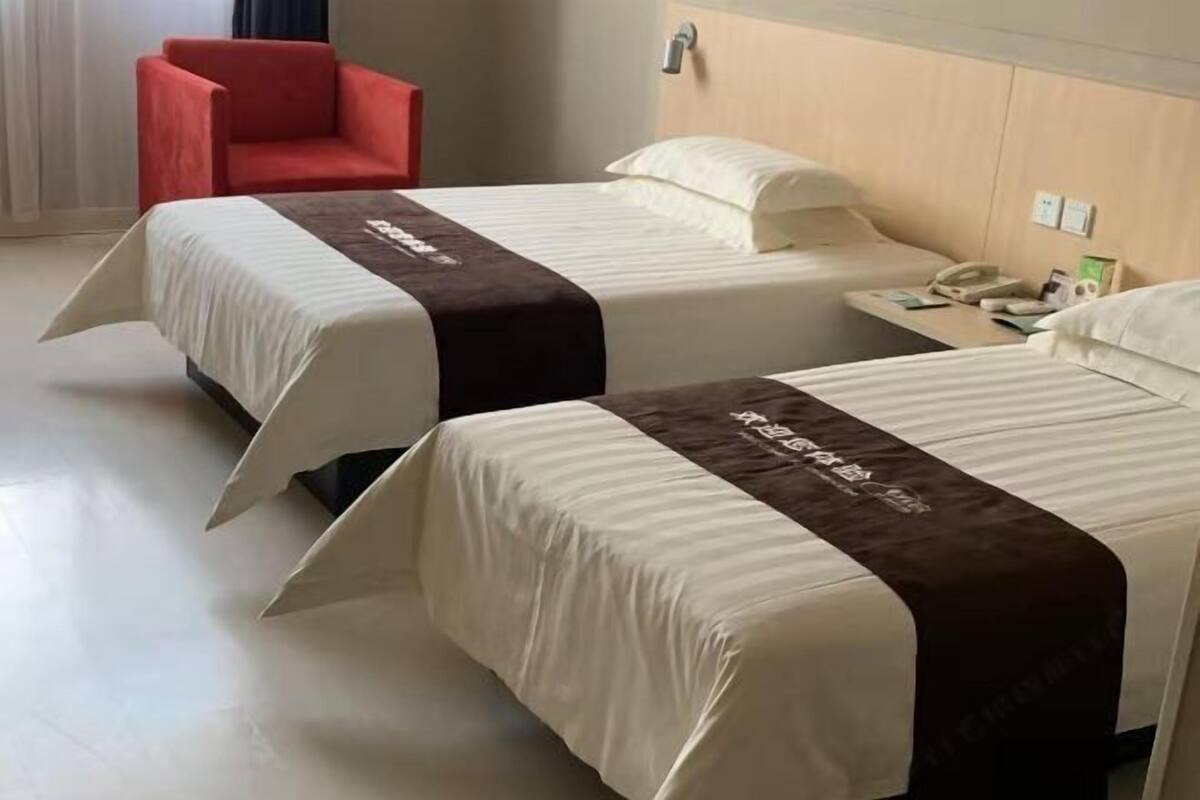 Two beds with white and brown bedspreads, a red chair, a bedside table with a phone, and a wall-mounted light.
