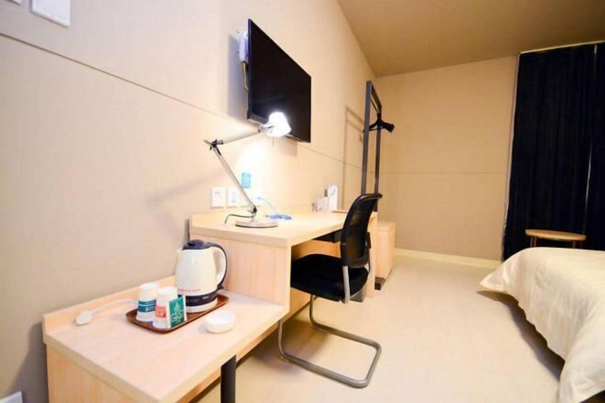 A hotel room with a desk, chair, bed, TV, and a small table with a kettle and cups.