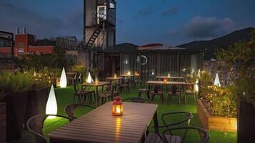 An outdoor dining area with tables and chairs, a wooden structure, and a view of the cityscape.