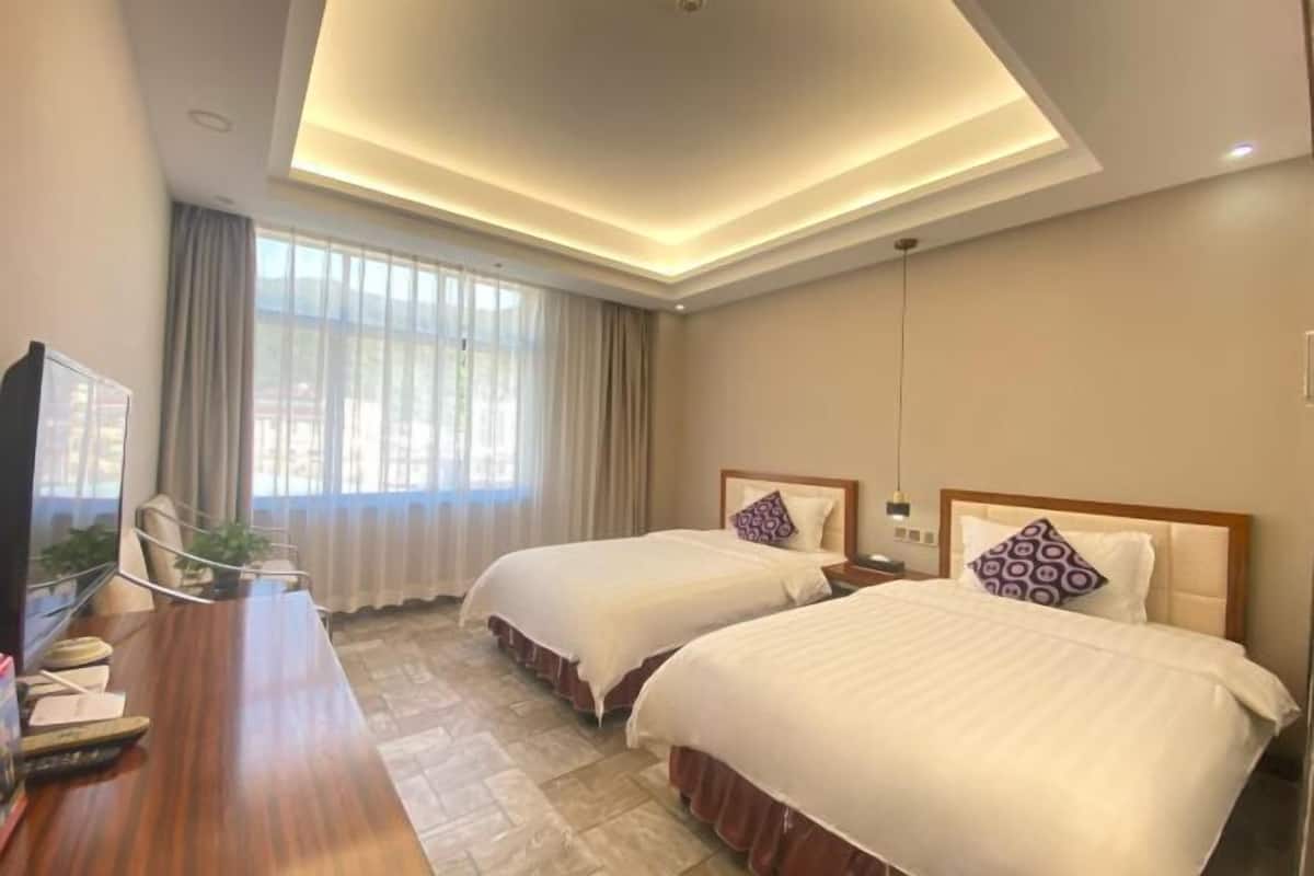 A hotel room with two beds, a wooden desk with a television, and a large window with curtains.