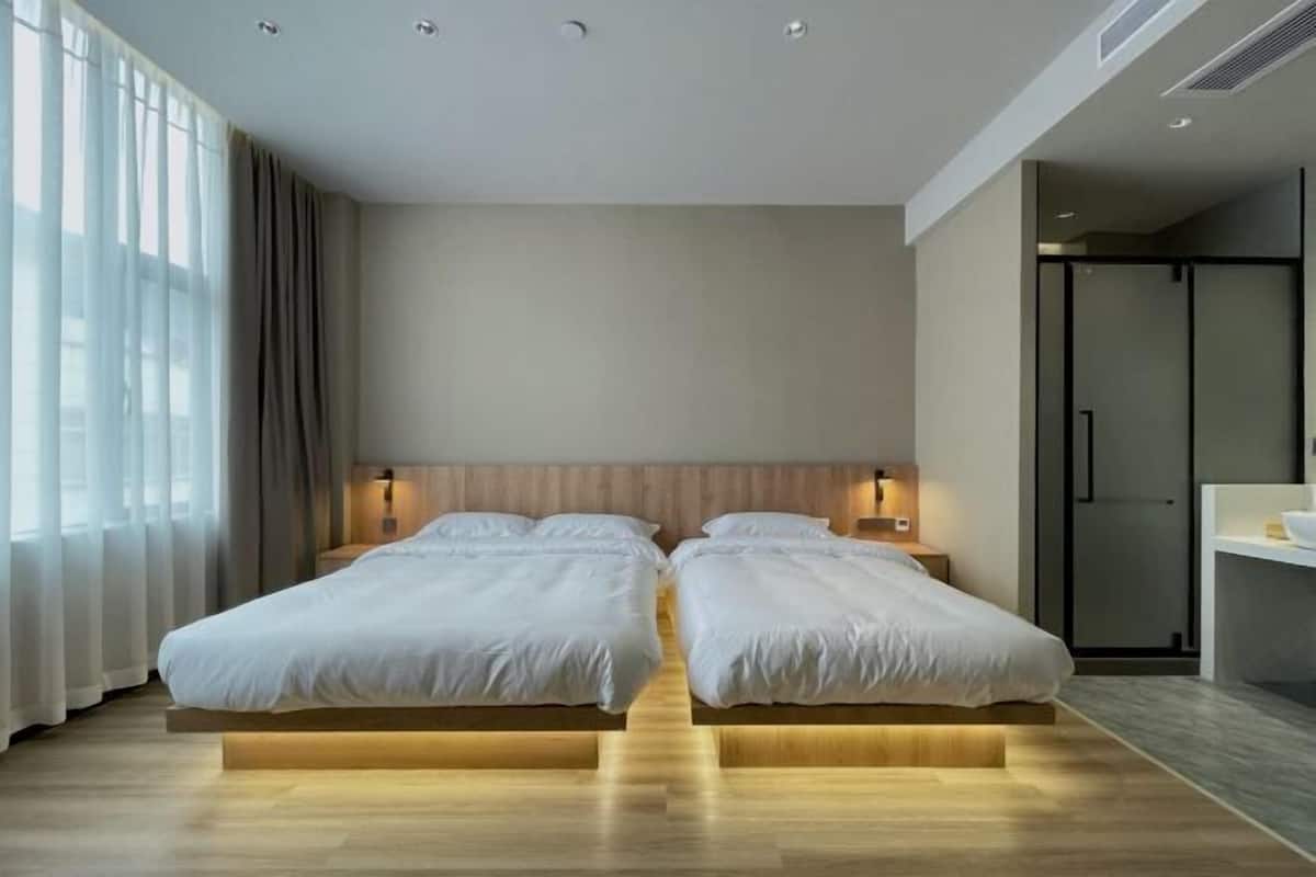 A modern hotel room with two beds, a wooden headboard, bedside lamps, a large window with curtains, and a walk-in closet.