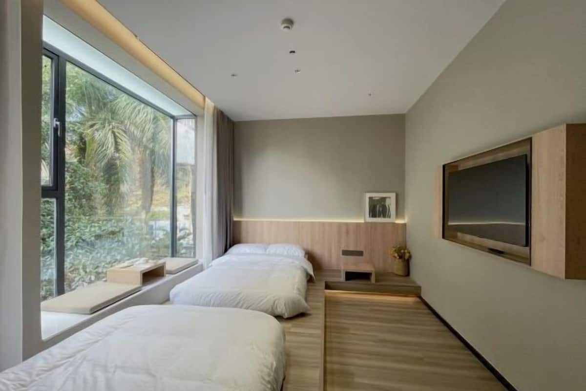 A modern hotel room with two beds, a large window, a wooden floor, and a mounted TV.