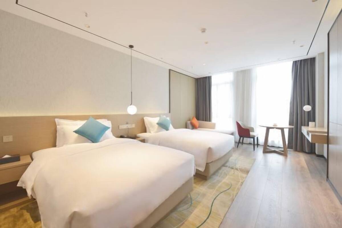 Ein Hotelzimmer mit zwei Betten, einem kleinen Tisch, einem Stuhl und einem großen Fenster mit Vorhängen.