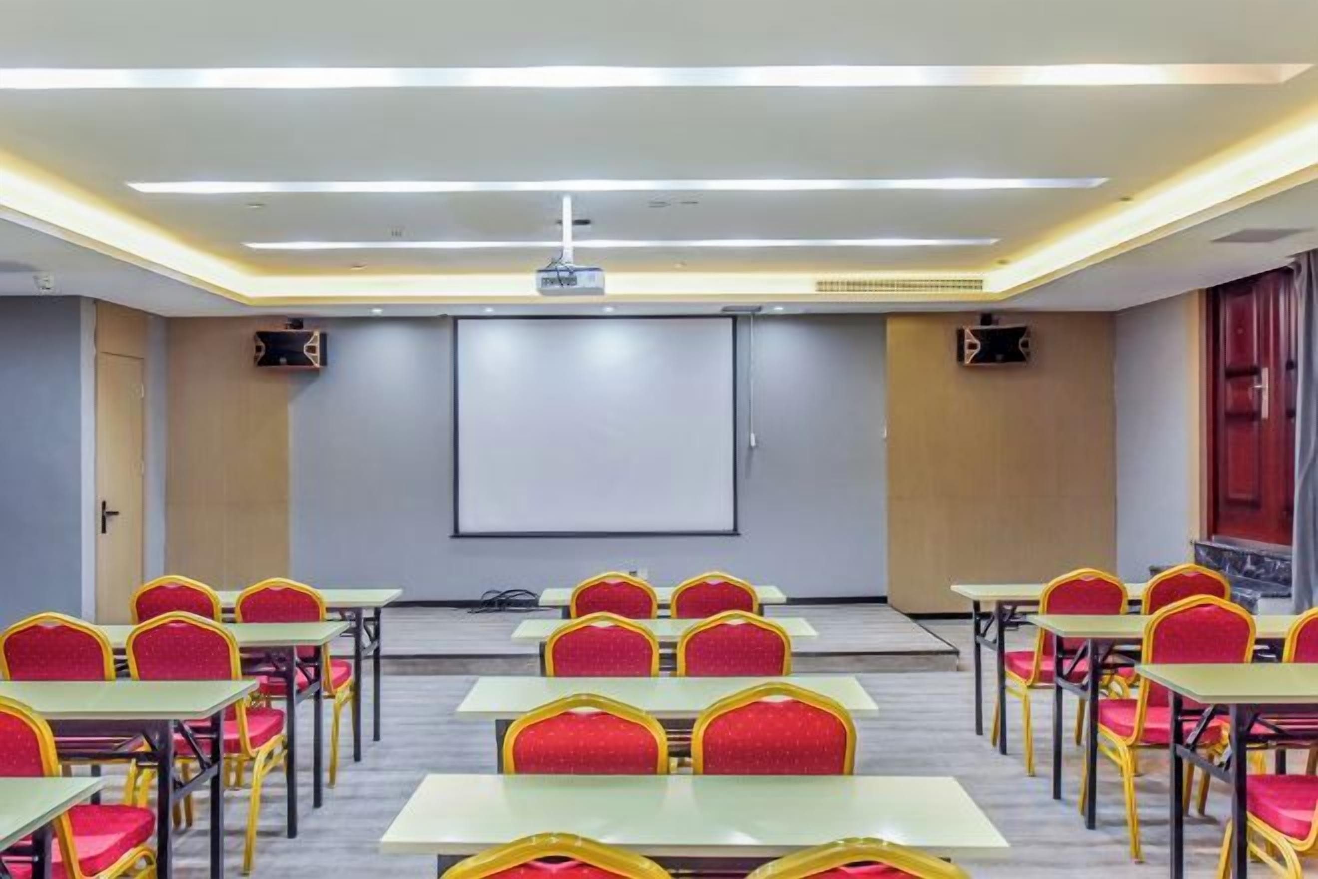 Un salón de conferencias con filas de mesas y sillas, una pantalla de proyector y una pantalla para presentaciones.