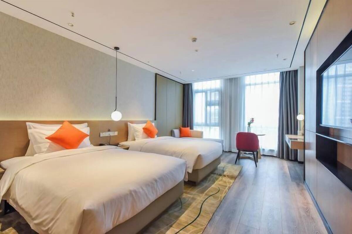 Ein Hotelzimmer mit zwei Betten, einem roten Sessel, einem Schreibtisch und großen Fenstern.