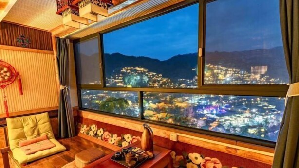 A room with a large window overlooking a cityscape at night, a wooden table with a tray of items, and a sofa with cushions. - Xijiang Qianhu Miaozhai Hanshe Inn (Observation Deck Store) (Leishan)