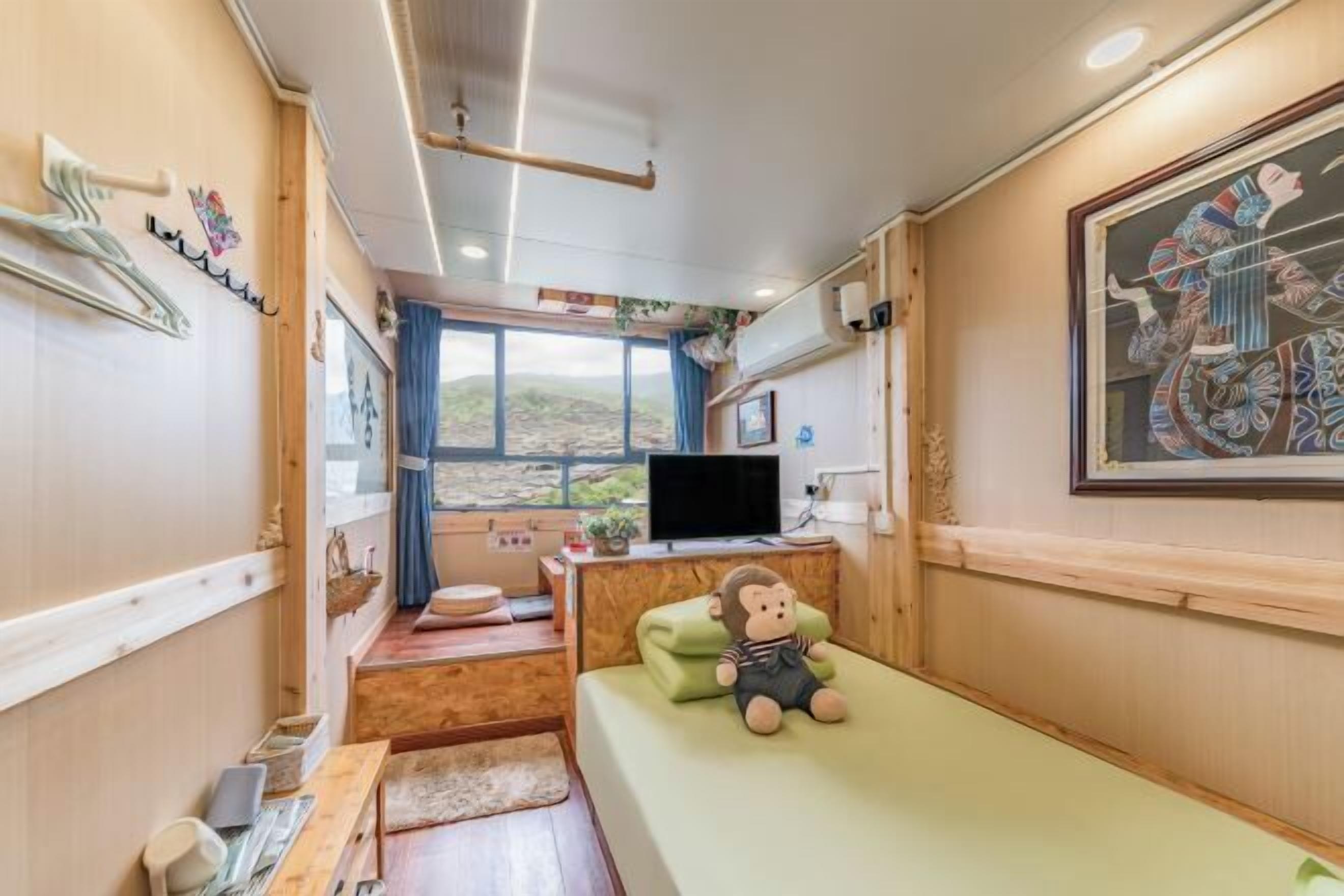 Quarto compacto com uma cama, uma escrivaninha com computador, uma janela com vista e um pequeno espaço para sentar.