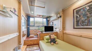 Quarto compacto com uma cama, uma escrivaninha com computador, uma janela com vista e um pequeno espaço para sentar.