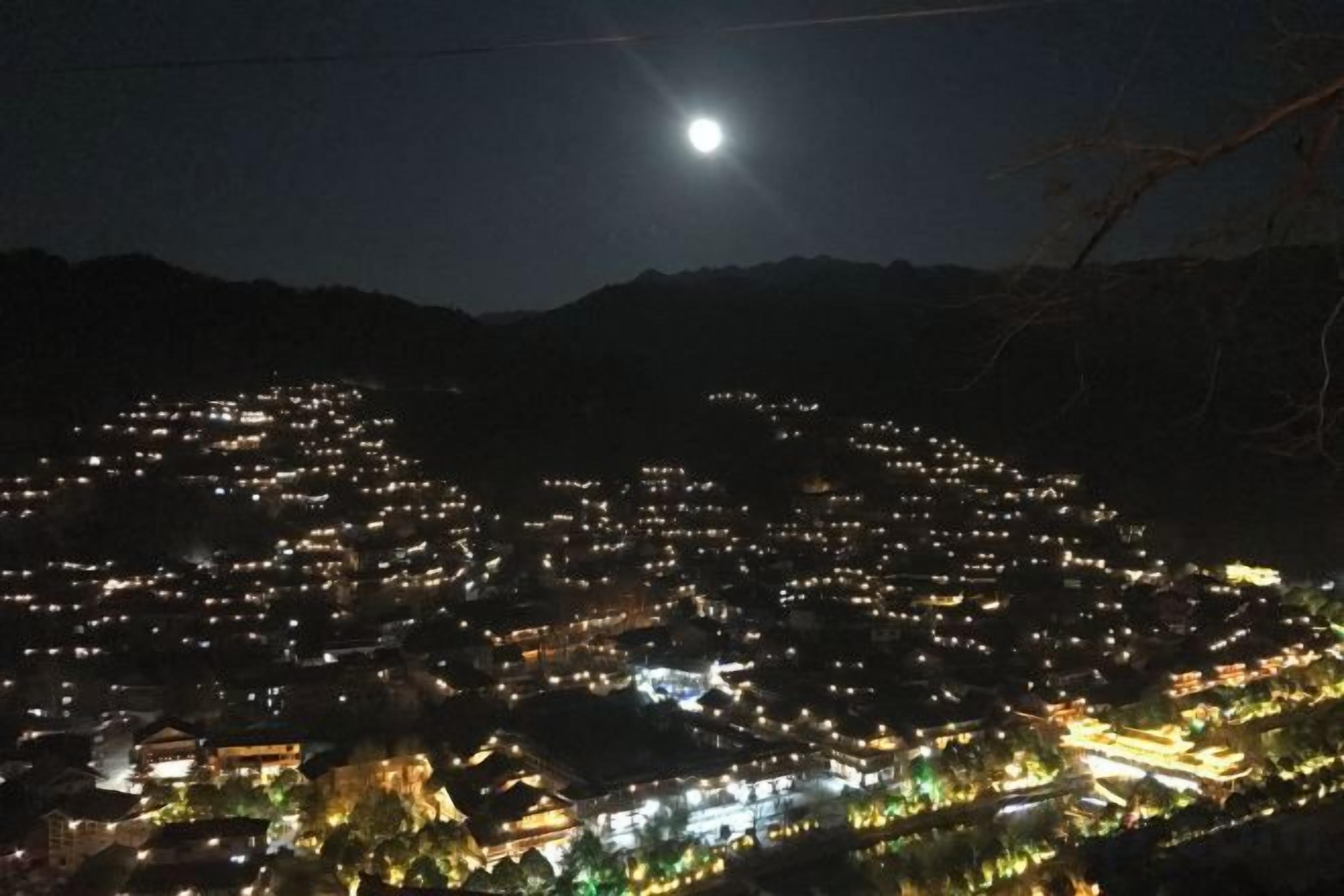 Une vue nocturne d’une ville, avec une colline imposante en arrière-plan et une lune bien visible.