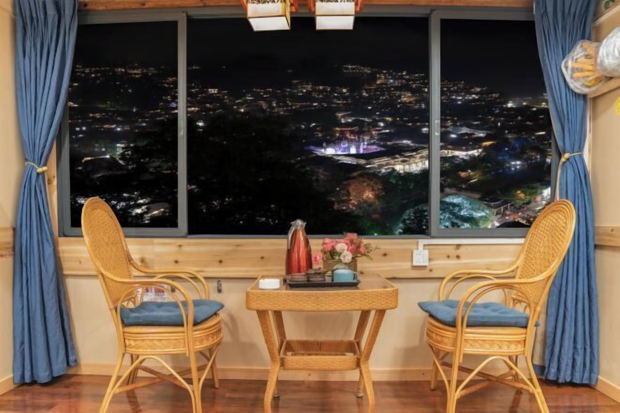 A room with a city view, two wicker chairs, a small table with a vase and a kettle, and curtains.