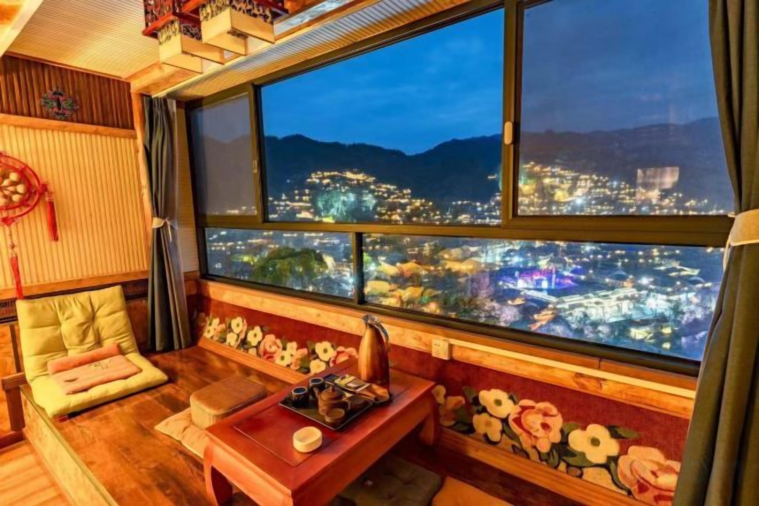 Habitación con una ventana grande que ofrece vistas de la ciudad de noche, un banco de madera y una pequeña mesa con un adorno.