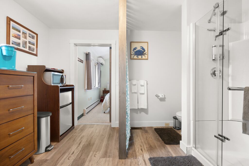 A modern bathroom with a glass shower enclosure, a wooden dresser, and a visible hallway leading to a bedroom.