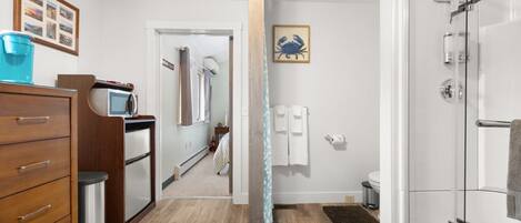 A modern bathroom with a glass shower enclosure, a wooden dresser, and a visible hallway leading to a bedroom.