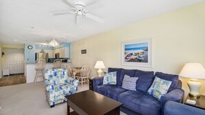 Sala de estar com sofá azul, poltrona estampada e mesa de centro. Há uma área de jantar com cadeiras e mesa, uma cozinha com armários e eletrodomésticos, e um ventilador de teto.