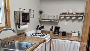 Une cuisine compacte dotée d’un plan de travail en granit, d’un évier en acier inoxydable, de rideaux blancs, d’une cuisinière à gaz et d’une machine à café.