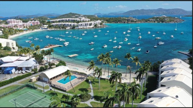 Un hotel costero con varias edificaciones, un puerto deportivo con barcos, una piscina y palmeras.