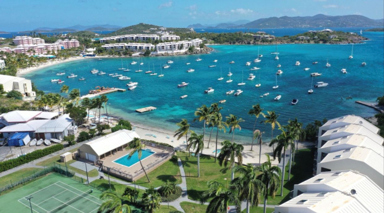 A coastal hotel with multiple buildings, a marina with boats, a swimming pool, and palm trees.