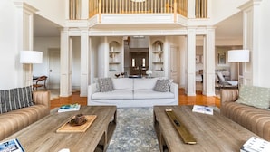 Un vaste espace de vie doté d’un haut plafond, d’un canapé blanc central, de deux tables basses en bois et d’un balcon avec une balustrade.