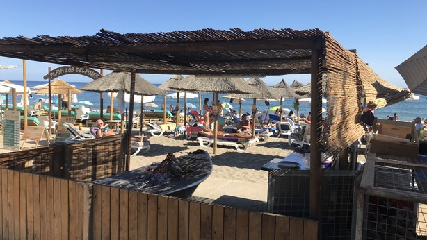 Un área junto a la playa con estructuras de madera, sombrillas, una parrilla y sillones de descanso.