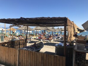 Eine Strandlage mit Holzkonstruktionen und Sonnenschirmen, einem Grill und Liegestühlen.