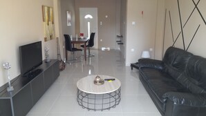 Een moderne woonkamer met een flatscreen televisie, een ronde salontafel, een zwarte leren bank en een eethoek op de achtergrond.