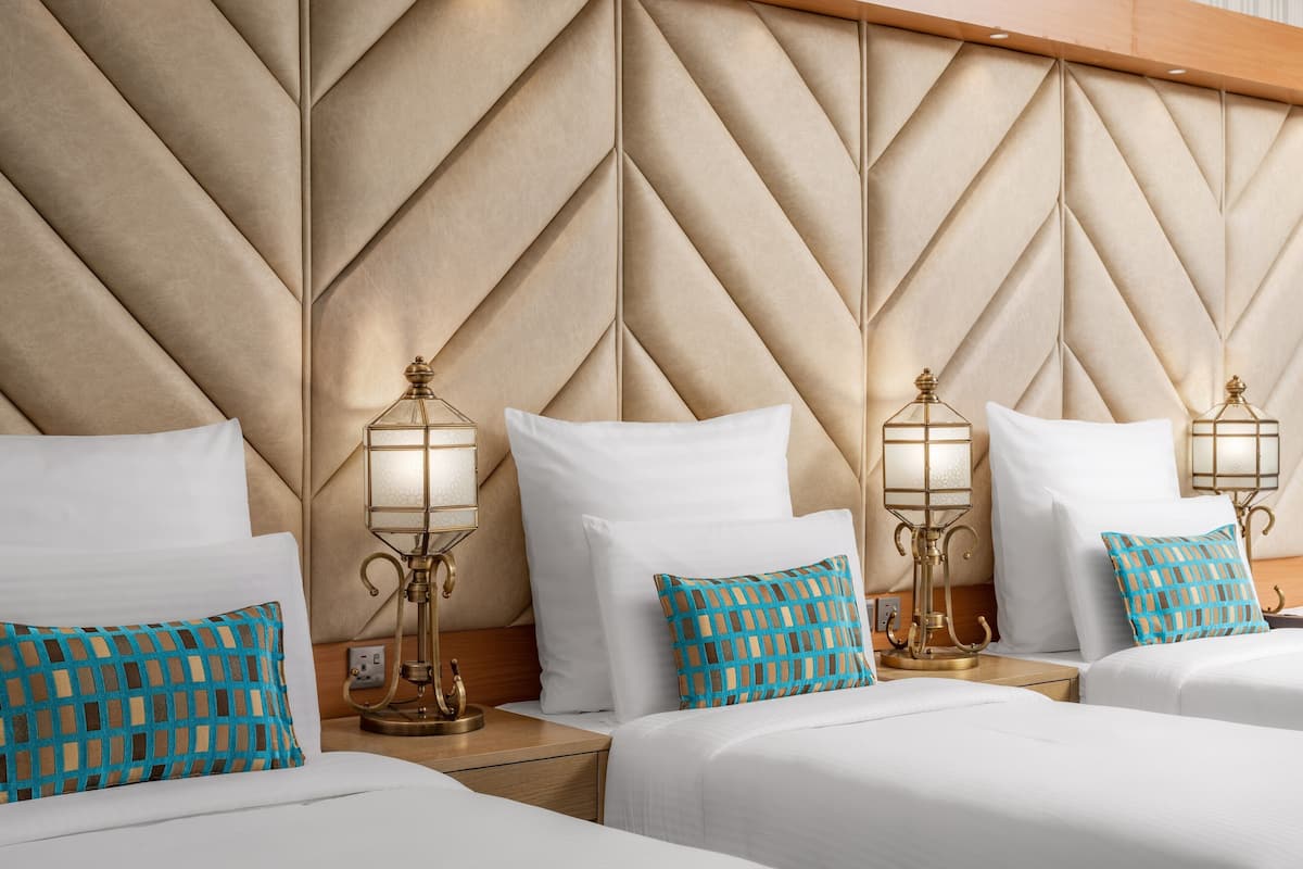 Une chambre d’hôtel dotée d’une grande tête de lit à motifs, de deux tables de chevet avec des lampes et de deux lits avec du linge de lit blanc et des oreillers à motifs.