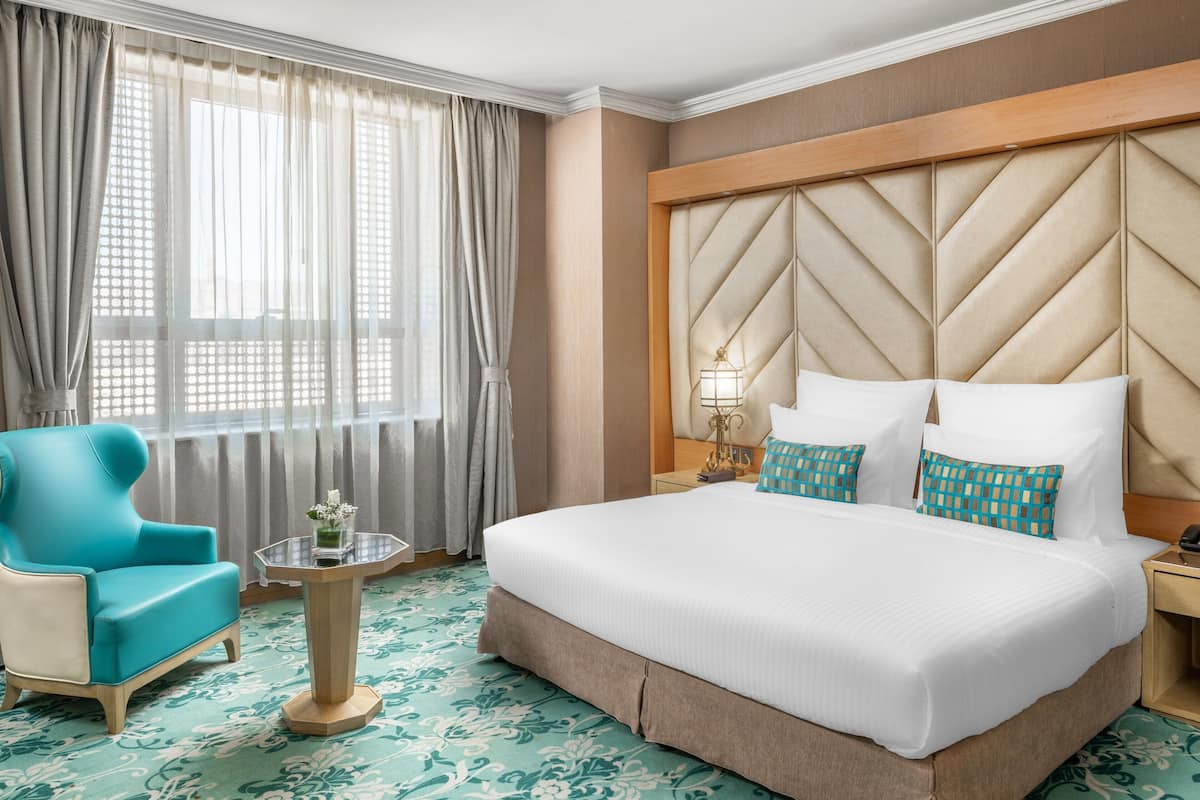 Une chambre d’hôtel avec un grand lit, un fauteuil bleu, une petite table avec un vase et une fenêtre avec des rideaux.