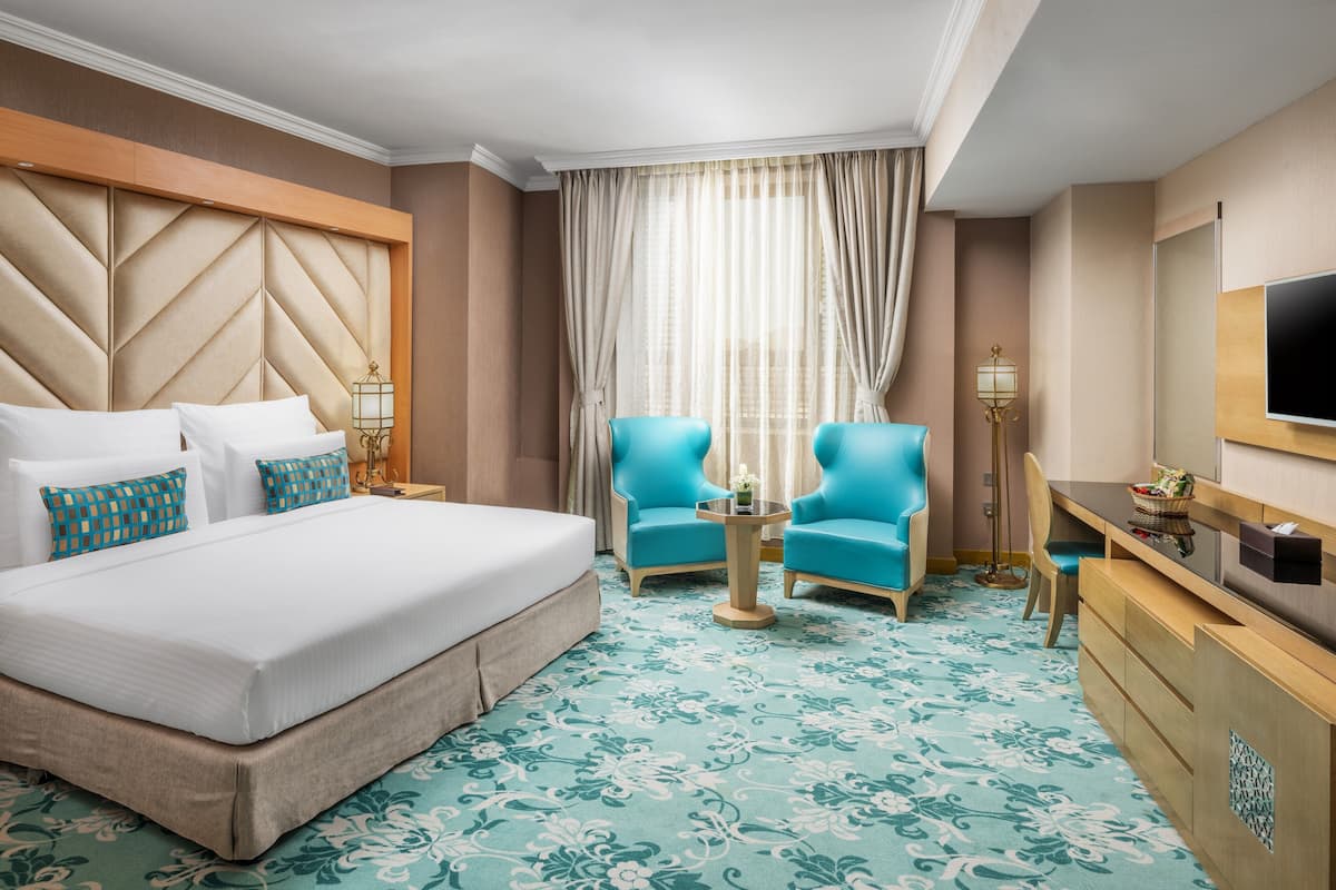 Une chambre d’hôtel avec un grand lit, deux fauteuils bleus, une petite table, un bureau avec une télévision et une grande fenêtre avec des rideaux.